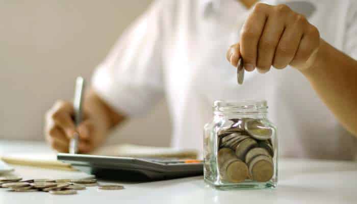 How can fd rates influence your deposit planning 1 | tycoonstory media Person placing a coin into a glass jar full of coins while budgeting at a desk.