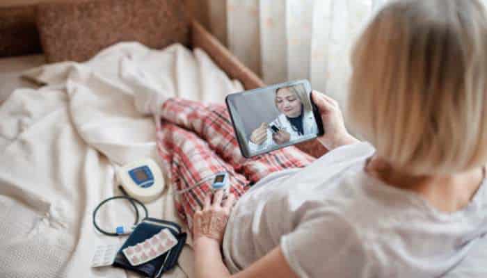How remote patient monitoring is improving home healthcare in ontario 1 | tycoonstory media Woman in bed holding a tablet with a doctor on video call; home medical devices and a pill pack are nearby.