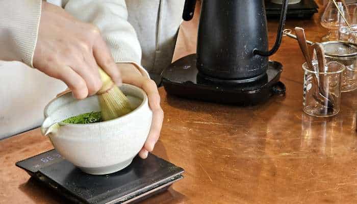 How to use a bamboo whisk to achieve the perfect froth on ceremonial grade matcha 1 | tycoonstory media Person preparing matcha tea using a traditional bamboo whisk in a bowl on a kitchen counter.