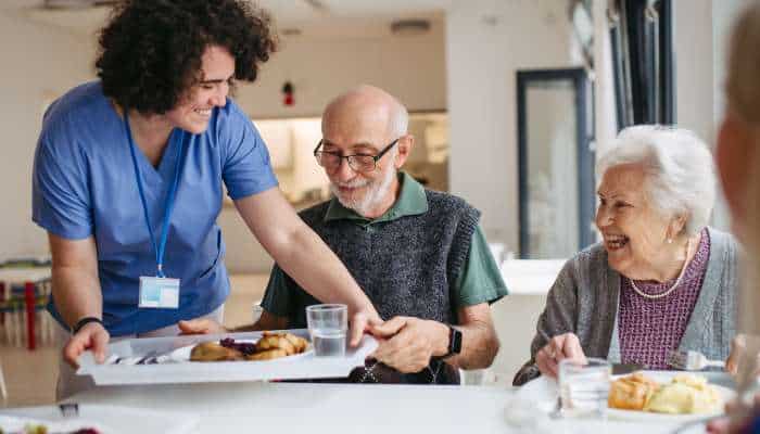 Include medical and health information | tycoonstory media Caregiver in blue scrubs serving a meal to two smiling elderly residents at a bright dining table.