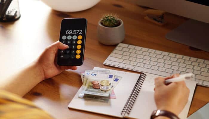 Why tracking fees in real time protects your margins - tycoonstory | tycoonstory media Person writes in a notebook at a wooden desk with euros and coins; a smartphone shows 69,572 on a calculator app.