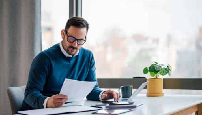 Nre account vs nro account 1 | tycoonstory media Man with glasses in a blue sweater sits at a bright desk, reviewing papers beside a laptop and cup. A small potted plant sits by the window.