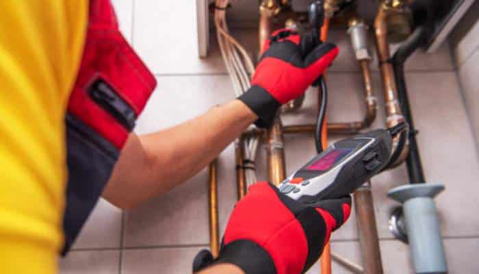 _ signs your sewer line may have a hidden leak 1 | tycoonstory media Plumber in a yellow shirt and red gloves uses a handheld meter to inspect copper pipes under a sink.
