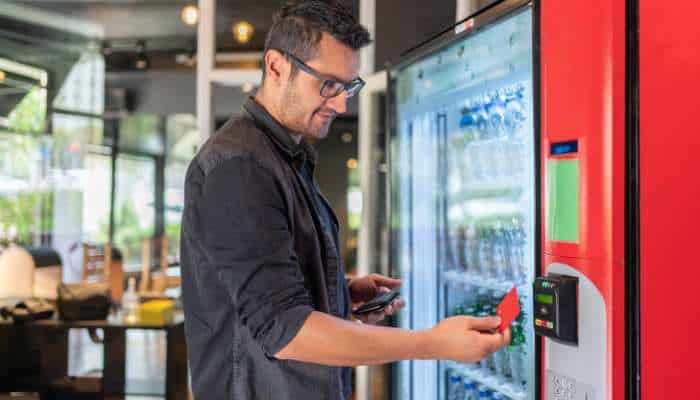 Smart vending machines 1 | tycoonstory media Man with glasses uses a red vending fridge, holding a card and a smartphone to pay.