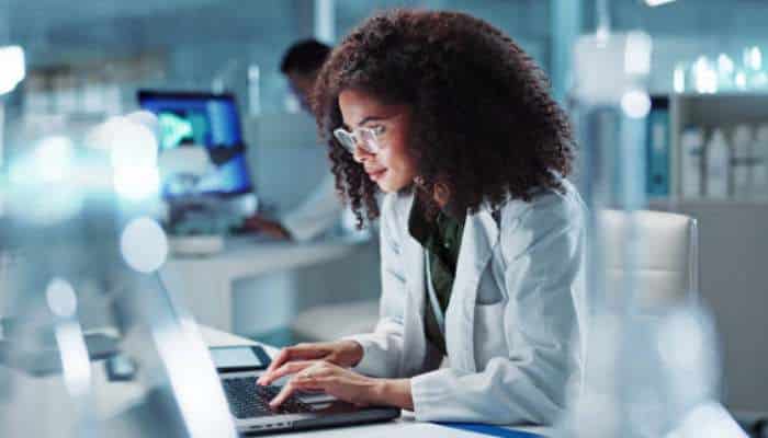The attribution problem almost every healthcare brand faces 1 | tycoonstory media Scientist in a white lab coat typing on a laptop in a bright, modern laboratory.