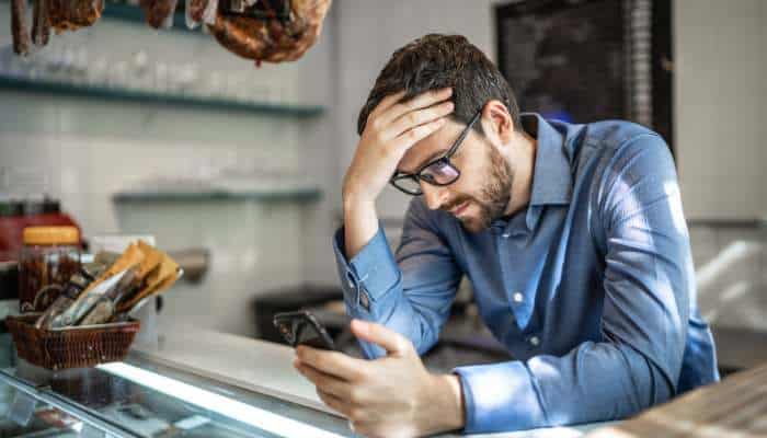 The payment problem every small org ignores too long 1 | tycoonstory media Man with glasses at a deli counter, looking at his phone with one hand on his forehead.