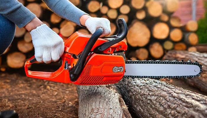 Practical tips for maximizing cutting capacity safely - tycoonstory | tycoonstory media Person wearing white gloves operating an orange chainsaw to cut through a log outdoors, with a stack of cut logs in the background.