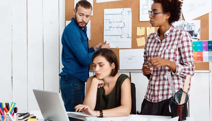 Expecting the first release to do everything - tycoonstory | tycoonstory media Three coworkers collaborating around a laptop at a bright office desk; a seated woman types while two colleagues discuss near a corkboard and charts.