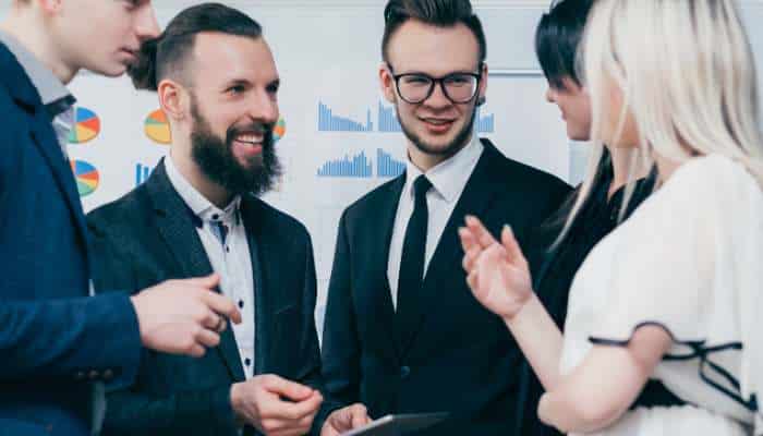 What makes a strong employee experience strategy 1 | tycoonstory media Group of four professionals in suits discussing and smiling around a tablet, with charts on the wall behind them.