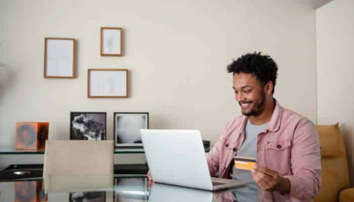Why businesses need a current account for daily transactions 1 | tycoonstory media Smiling man using a laptop at a glass desk while holding a credit card for online shopping.
