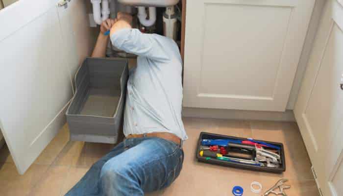 Why diy plumbing fixes often lead to bigger problems 1 | tycoonstory media Person lying on the floor under a kitchen sink, repairing pipes with a toolkit nearby.