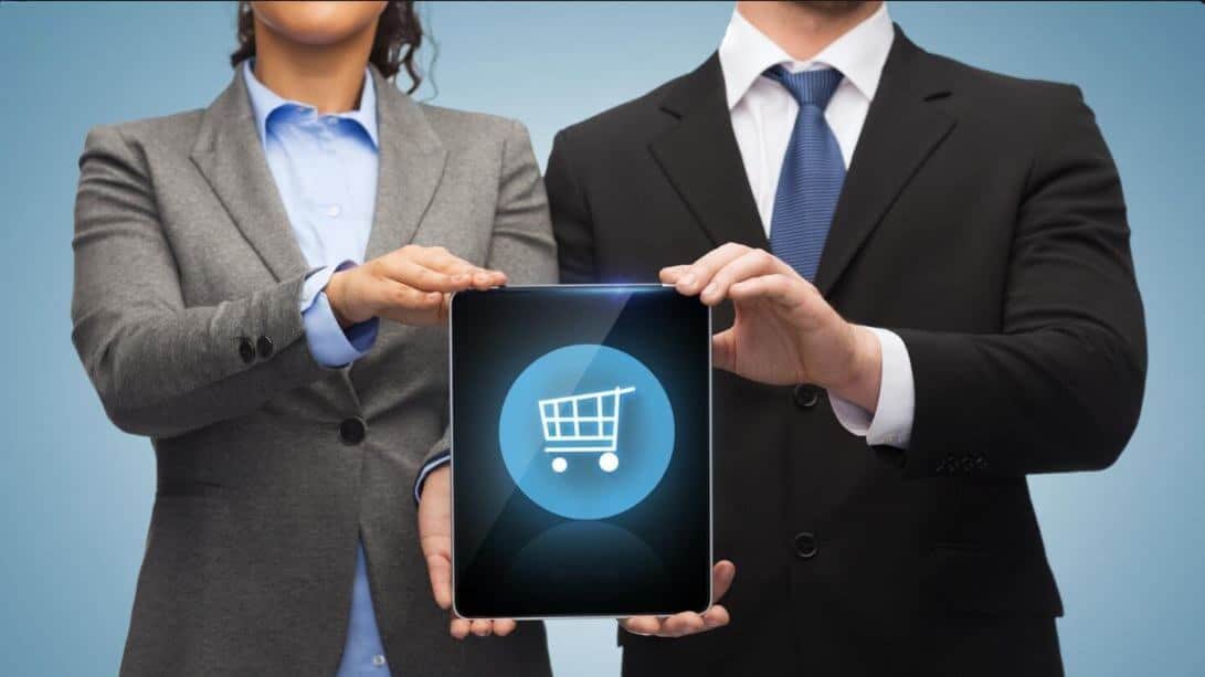 Compete on the relationship, not the transaction - tycoonstory | tycoonstory media Two professionals in suits hold a tablet showing a blue shopping cart icon, against a blue background.