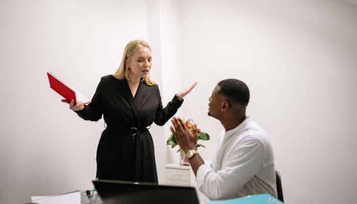 Workplace harassment 1 | tycoonstory media Woman in a black dress stands by a desk, holding a red folder and gesturing toward a seated man who clasps his hands in front of him.