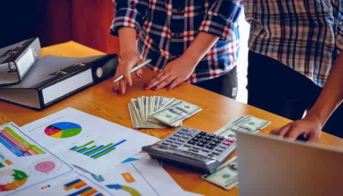 Mastering cash flow and capital allocation - tycoonstory | tycoonstory media Two people budgeting at a wooden table with charts, graphs, a calculator, folders, laptop, and cash on the desk.