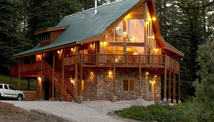 Build energy performance into the architecture - tycoonstory | tycoonstory media Contemporary log cabin featuring a steep metal roof, expansive glass windows, natural log walls, stone foundation, and wraparound wooden deck surrounded by dense forest.