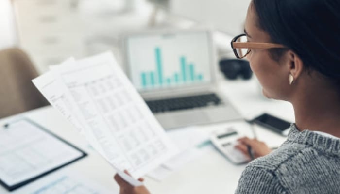 Building a smarter employee expense process | tycoonstory media Analyst with glasses reviewing printed financial documents while a laptop shows bar charts in the background