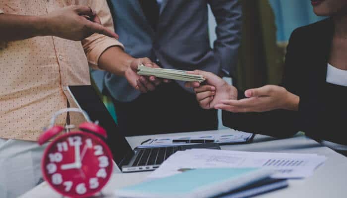 Financial commitments that continue long after you move in | tycoonstory media Two hands exchanging a stack of cash across a desk with papers, laptop, and a pink alarm clock in view.
