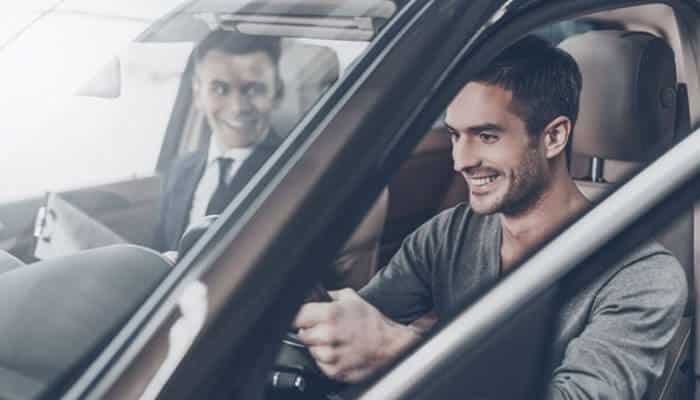 | tycoonstory media Man smiling while driving a car, with a suited colleague visible in the back seat through the window in a showroom-like setting.