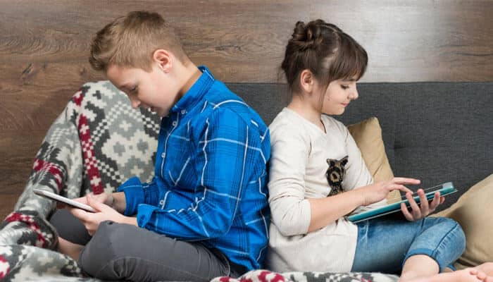 A language kept alive at dinner volume - tycoonstory | tycoonstory media Two children sit back-to-back on a sofa, each absorbed with a tablet. A cozy blanket covers the armrest nearby.