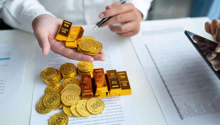 Whythepriceyou payfor goldmatters - tycoonstory | tycoonstory media Hands handling gold coins and bars over financial documents on a desk, with a pen nearby and a smartphone visible in the background.