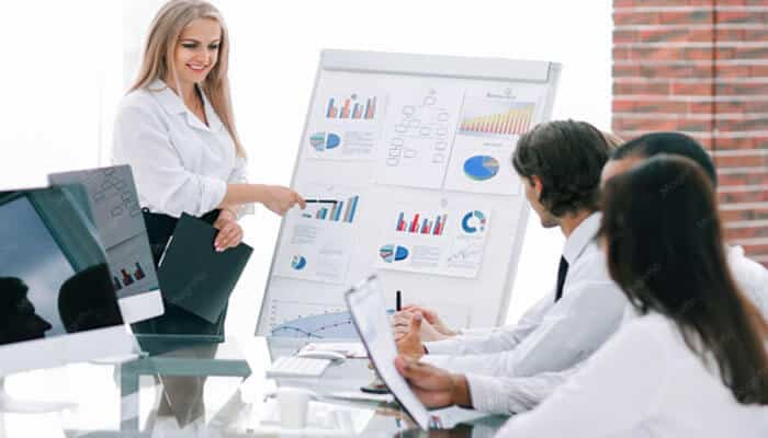 What to look for in a dynamics 365 partner | tycoonstory media Businesswoman presents a large chart board to a team during a meeting, with graphs and charts visible on the board.