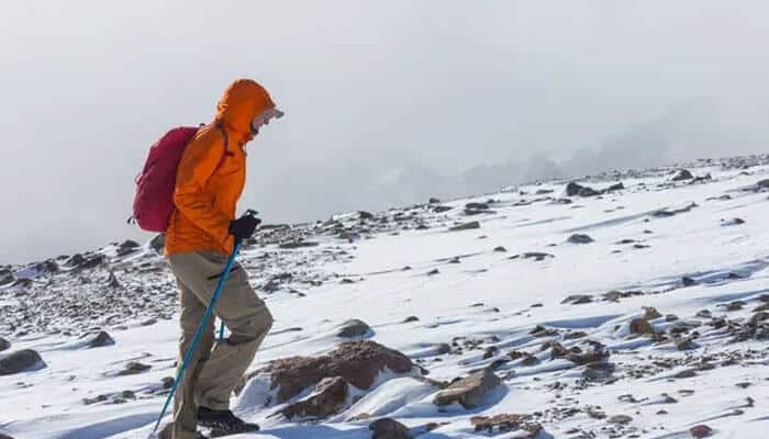 Altitude sickness medication: what to know - tycoonstory | tycoonstory media Hiker in an orange jacket and pink backpack uses trekking poles on a snowy, rocky mountain slope.