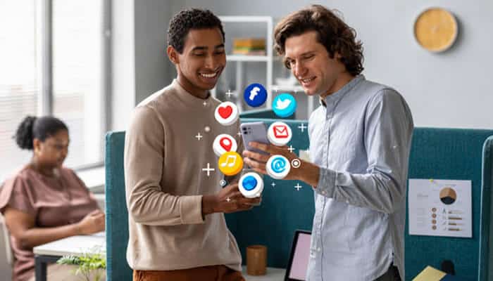 They streamline collaboration with the help of social media management tools - tycoonstory | tycoonstory media Two men standing in an office, looking at a smartphone with colorful social media icons floating around.
