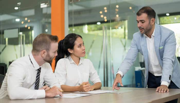 Give employees more choice over where they work - tycoonstory | tycoonstory media Three professionals in a conference room: a standing man points at documents on the table as two colleagues listen attentively.