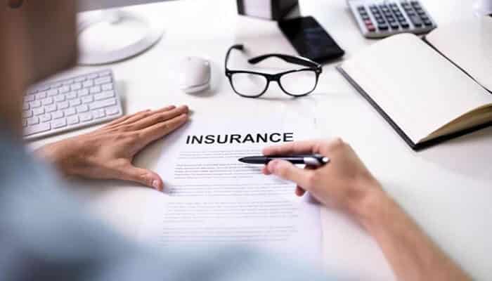 Insurance coverage can still matter - tycoonstory | tycoonstory media Person signing an insurance document at a white desk, with glasses, a notebook, calculator, and keyboard nearby.