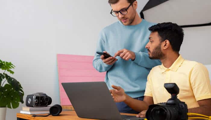 What founders should check before hiring locally - tycoonstory | tycoonstory media Two men collaborate at a desk with a laptop and camera gear, one showing something on a phone to the other.