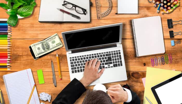 The budget line items that actually move - tycoonstory | tycoonstory media Top-down view of a person typing on a silver laptop at a cluttered wooden desk with notebooks, pencils, a plant, money, and office supplies.