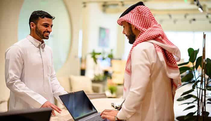 Top companies in the saudi arabia cloud and digital transformation industry | tycoonstory media Two men in an office chat and smile while standing at a desk with a laptop between them.