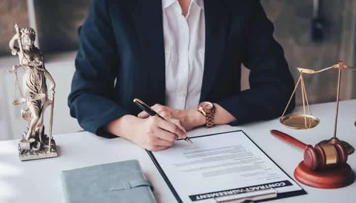 Urgent protective applications - tycoonstory | tycoonstory media Professional woman in a dark blazer signs a contract at a desk, with lady justice statue, scales, and a gavel nearby.