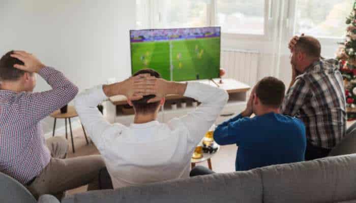 Streameast nfl 1 | tycoonstory media Four friends watching a soccer match on a tv in a living room, with one person on the left holding his head and others reacting in suspense, a christmas tree visible to the side.