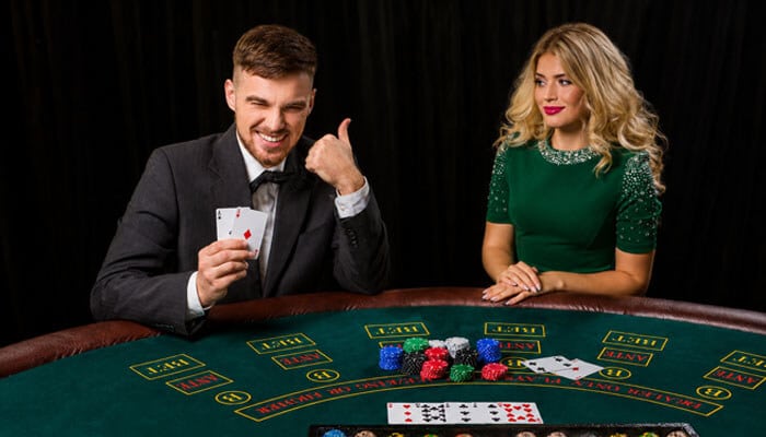 | tycoonstory media Smiling man in a suit at a casino table, showing two cards and giving a thumbs-up while a woman in a green dress watches nearby.
