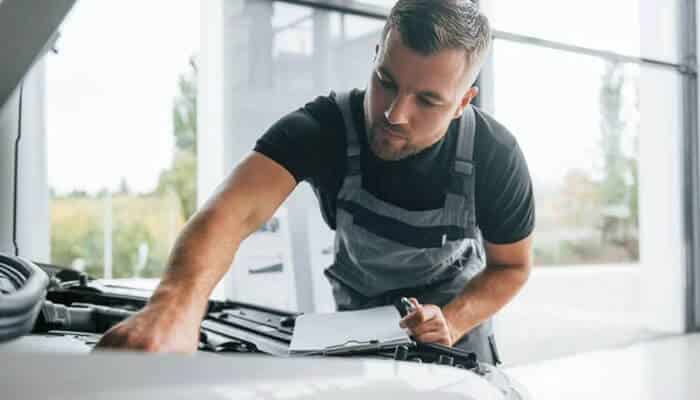 Autoshop solutions — best full-service automotive marketing agency | tycoonstory media Mechanic in gray overalls leaning over a car engine, holding a clipboard and inspecting under the hood.