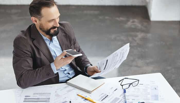 Texas has its own tax considerations | tycoonstory media Man in a brown blazer sits at a desk, looking at documents while checking a smartphone.