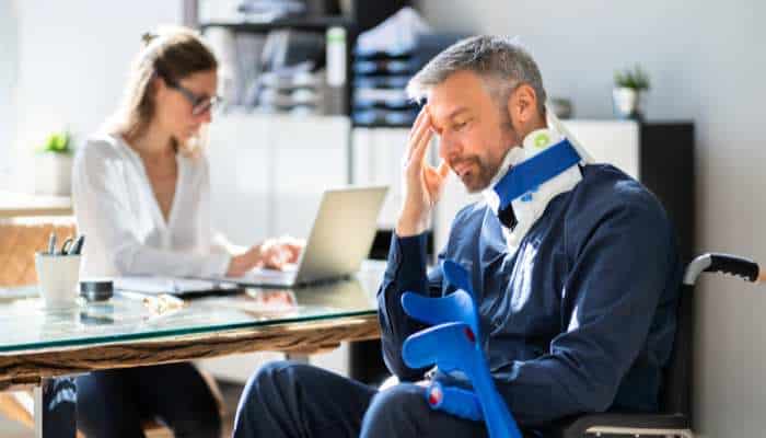 Why truck crash victims need legal help before evidence vanishes 1 | tycoonstory media Man in a wheelchair wearing a neck brace and arm sling sits at a desk in an office, head in hand, while a woman works on a laptop in the background.