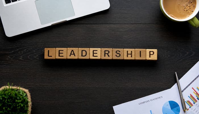 Philosophy of passionate leadership - tycoonstory | tycoonstory media Top-down view of wooden tiles spelling'LEADERSHIP' on a dark desk beside a laptop and a cup of coffee.