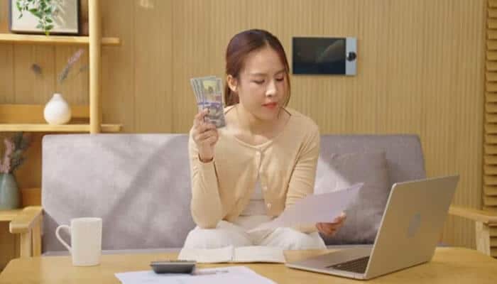 Senior and lead-level it salaries - tycoonstory | tycoonstory media Woman sitting on a sofa at a wooden desk, counting cash in one hand while reading a document with a laptop open nearby.