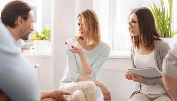 Reducing stigma in the workplace - tycoonstory | tycoonstory media Three colleagues in a bright office discuss; a woman holds a coffee cup as she speaks to the others in a casual meeting face to face.