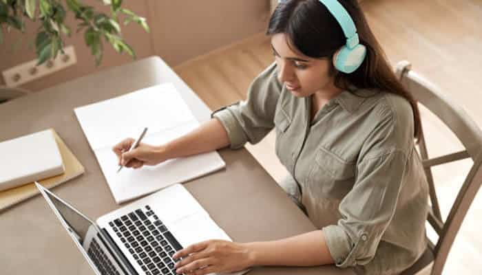 Go and look — that's the whole exercise | tycoonstory media Woman wearing turquoise headphones works on a laptop at a desk, taking notes on paper