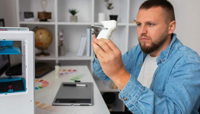 Consider processing and part design | tycoonstory media Man in a denim shirt inspecting a small white 3d-printed object beside a desktop 3d printer in a workshop
