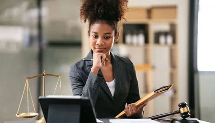 How representation protects a professional license | tycoonstory media Professional woman attorney sits at a desk with scales of justice and a gavel nearby, contemplating notes and documents in a law office.