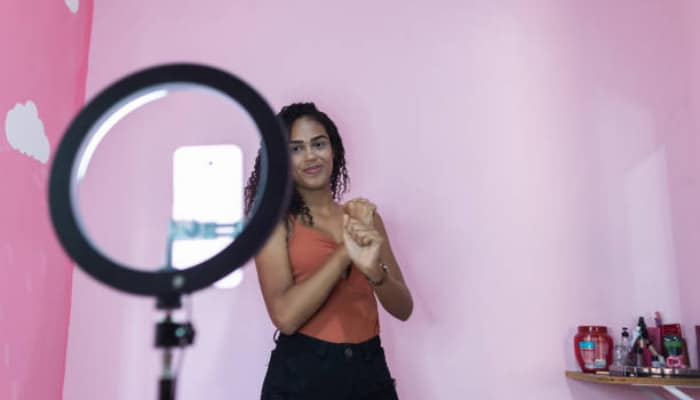 Build repeatable content series | tycoonstory media Woman in a peach tank top posing in a pink room; a ring light appears in the foreground with a smartphone on a stand nearby.