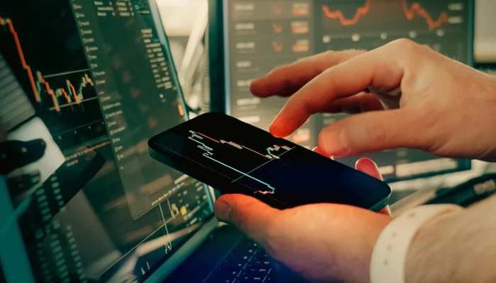 Can retail clients access institutional-grade forex pricing_ 1 | tycoonstory media Hands holding a smartphone displaying a stock price chart, with trading monitors and graphs in the background.