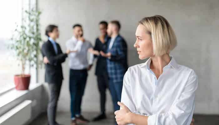 Workplace discrimination | tycoonstory media Woman in a white shirt stands in the foreground, looking to the right, as a group of coworkers chats in the background by a window.