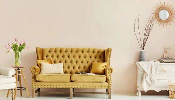 How to transition butter yellow into autumn | tycoonstory media Living room scene with a mustard-yellow tufted sofa, cream and yellow cushions, a small table with pink flowers, and a console table with a vase of branches and a sunburst mirror.