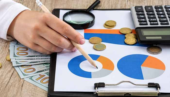 What the cost structure actually looks like | tycoonstory media Person marking financial charts on a clipboard with coins, cash, a calculator, and a magnifying glass nearby.