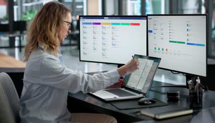Employee monitoring software 1 | tycoonstory media Professional woman at a desk uses a tablet while two monitors display colorful dashboards in the background.